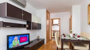A modern living room with a flat-screen TV, dark wooden cabinets, a dining area with a table set for two, and a hallway leading to a bedroom.