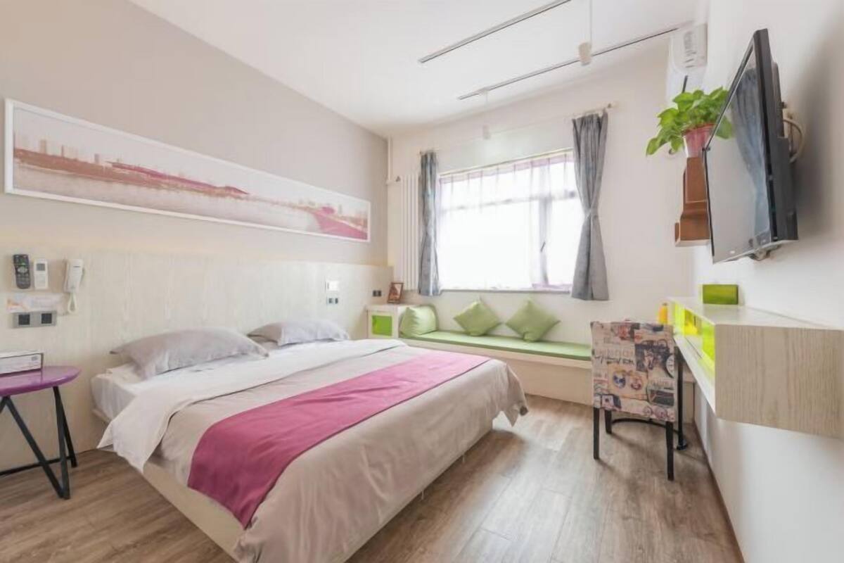 Une chambre d’hôtel comprenant un lit, un bureau, une chaise, une fenêtre avec des rideaux, un téléviseur fixé au mur et une plante fixée au mur.