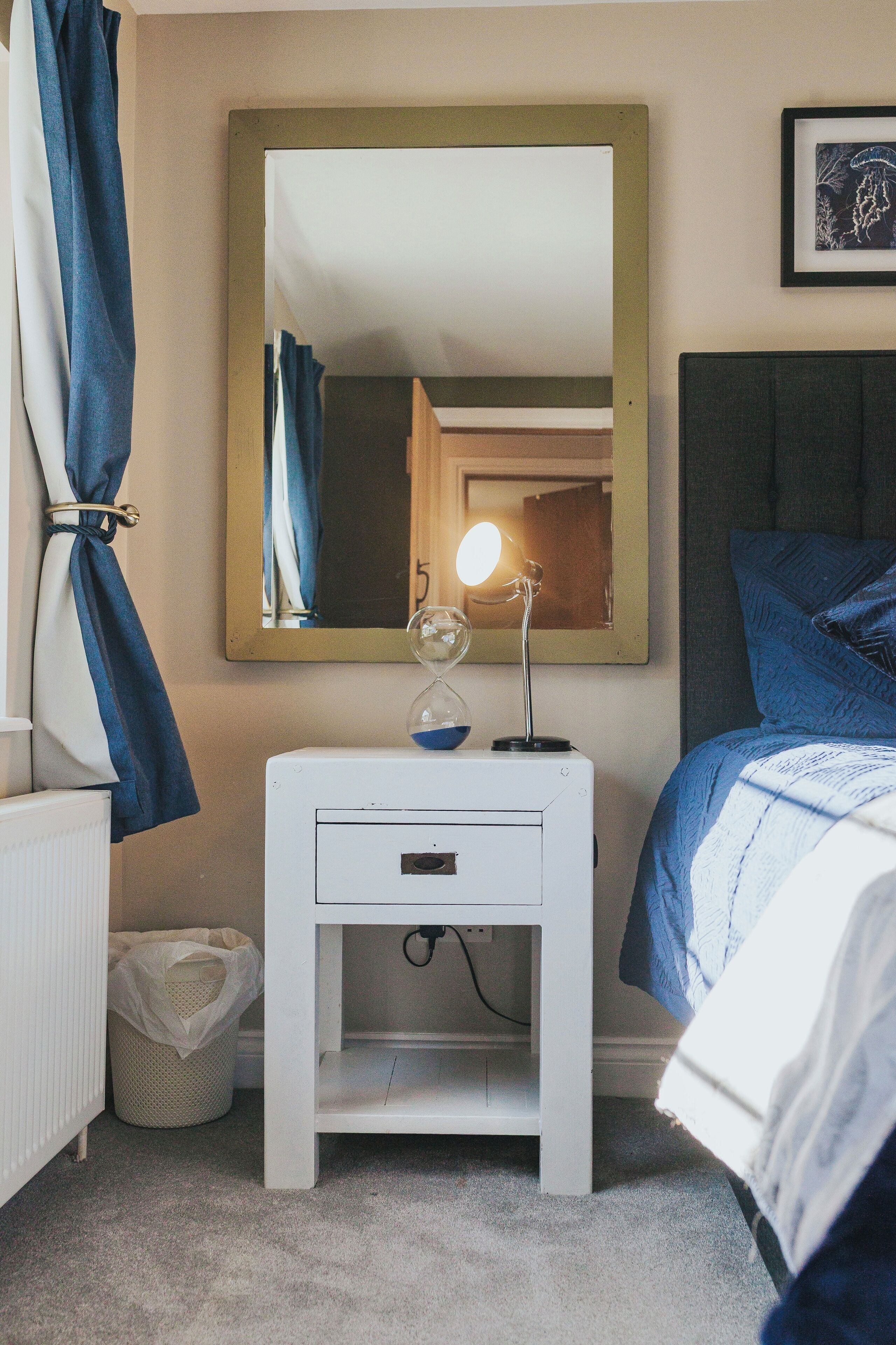Ein Schlafzimmer mit einem Bett, einem Nachttisch mit Lampe, einem Spiegel und einem gerahmten Bild an der Wand.