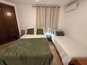 Une chambre avec un grand lit, deux petits lits, une armoire, un climatiseur et une table de chevet avec une lampe.