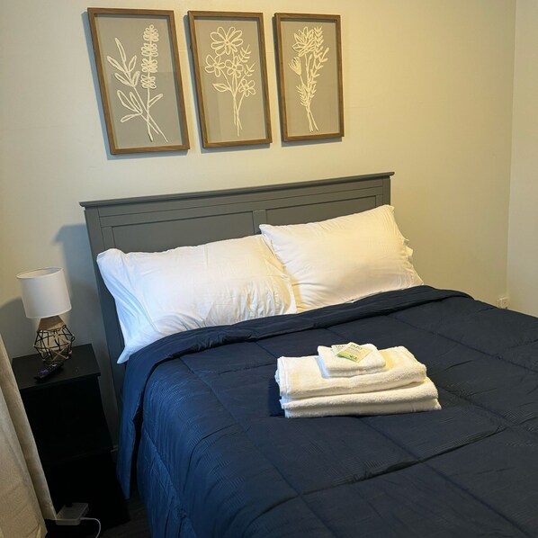 Una cama bien hecha con una colcha azul oscuro y almohadas blancas, una mesita de noche con una lámpara, y tres láminas enmarcadas con motivos botánicos en la pared.