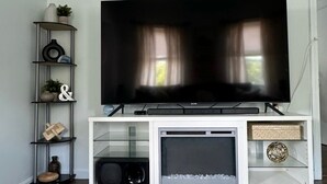 Een moderne woonkamer met een flatscreen televisie op een witte televisiemeubel, een glazen plank met decoratieve objecten, en een donkere houten vloer.
