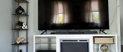 A modern living room with a flat-screen TV on a white entertainment unit, a glass shelf with decorative items, and a dark wooden floor.