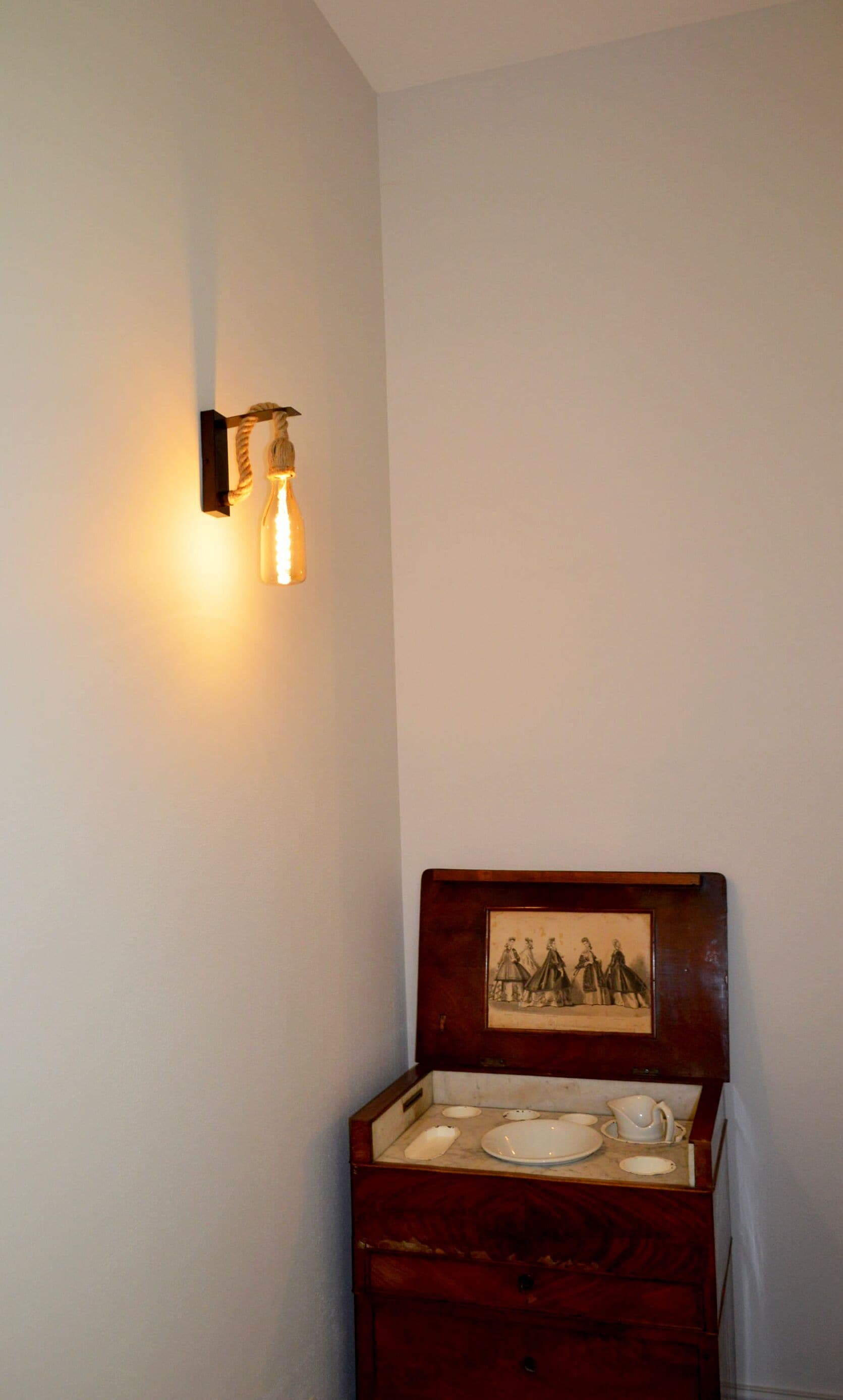 Un rincón con una lámpara de pared y un cofre de madera con un cuadro enmarcado y vajilla.