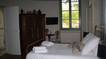 A neatly made bed with white linens and pillows, a wooden wardrobe, a small table with a chair, and a window with a view of greenery.
