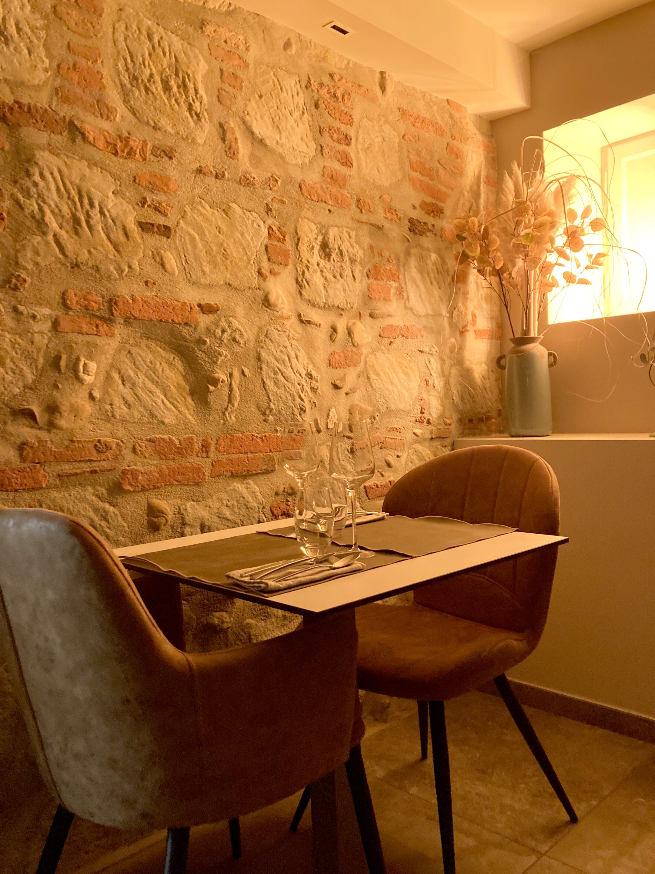 Un comedor con pared de ladrillo, dos sillas, una mesa con copas de vino y un florero con flores.