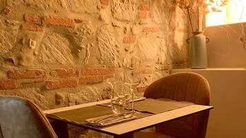 Un comedor con pared de ladrillo, dos sillas, una mesa con copas de vino y un florero con flores.