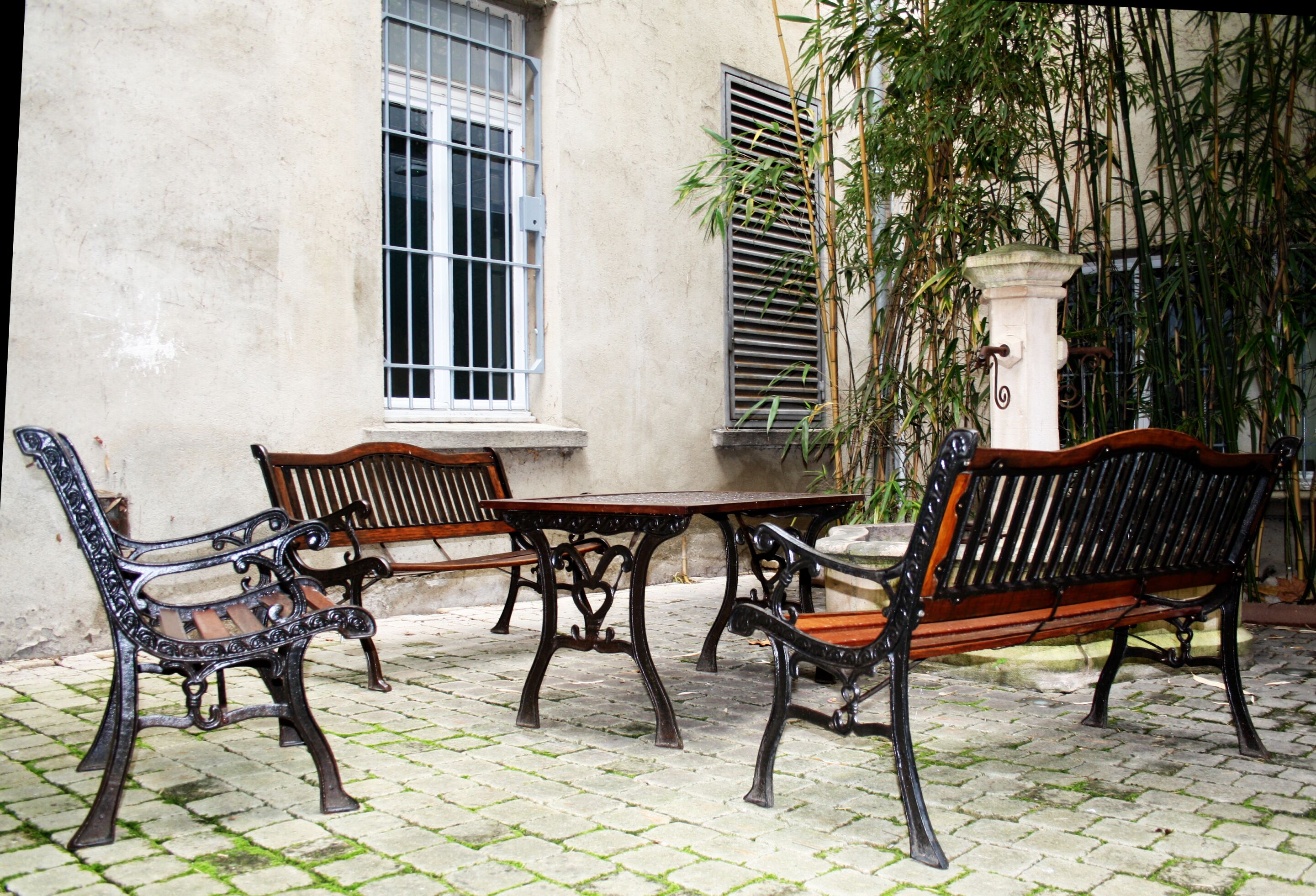 Une cour avec une table et trois bancs, une fenêtre avec des barreaux et un plant de bambou.
