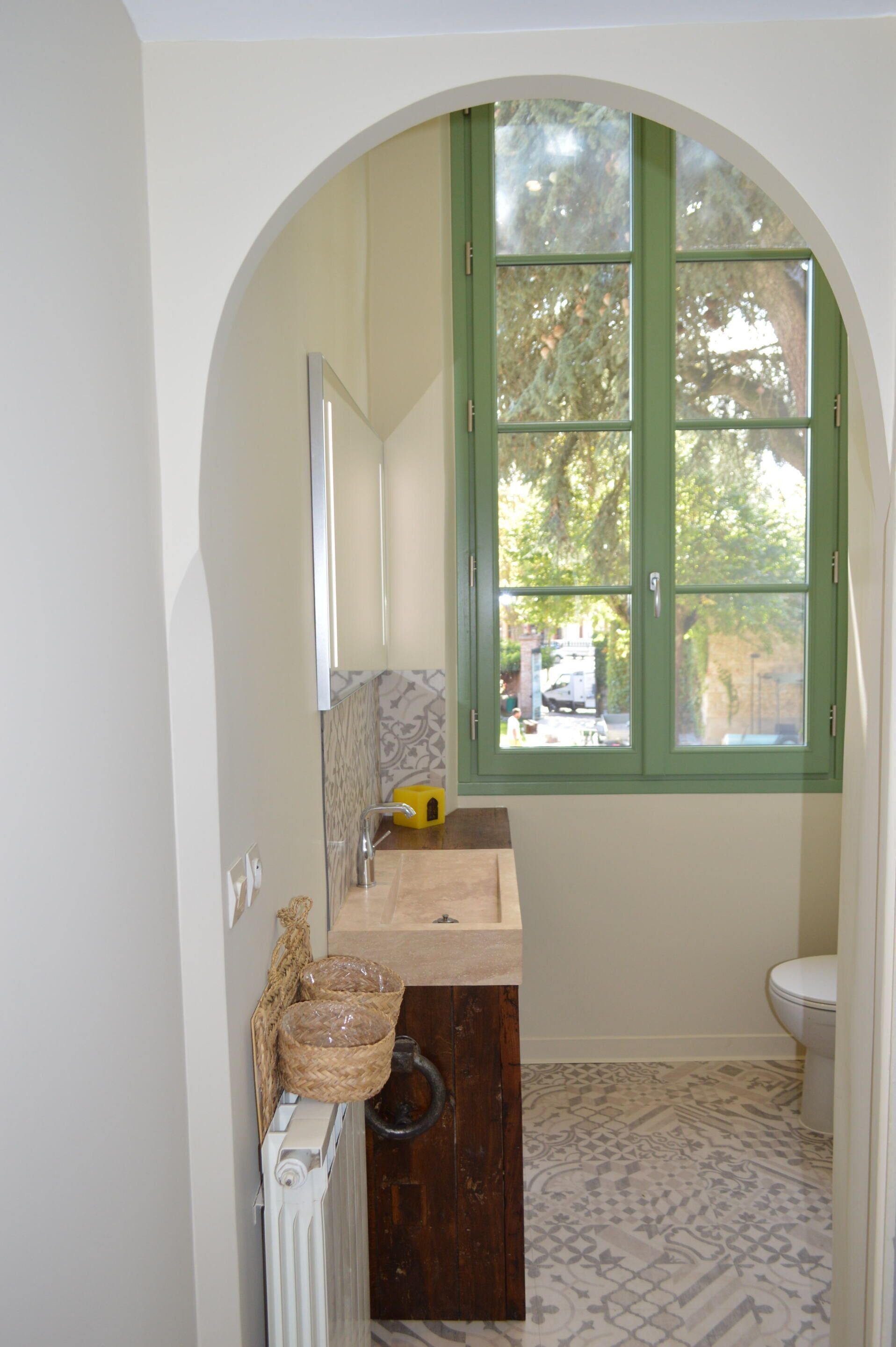 A bathroom with a white sink, a mirror, a window with green shutters, and patterned floor tiles.