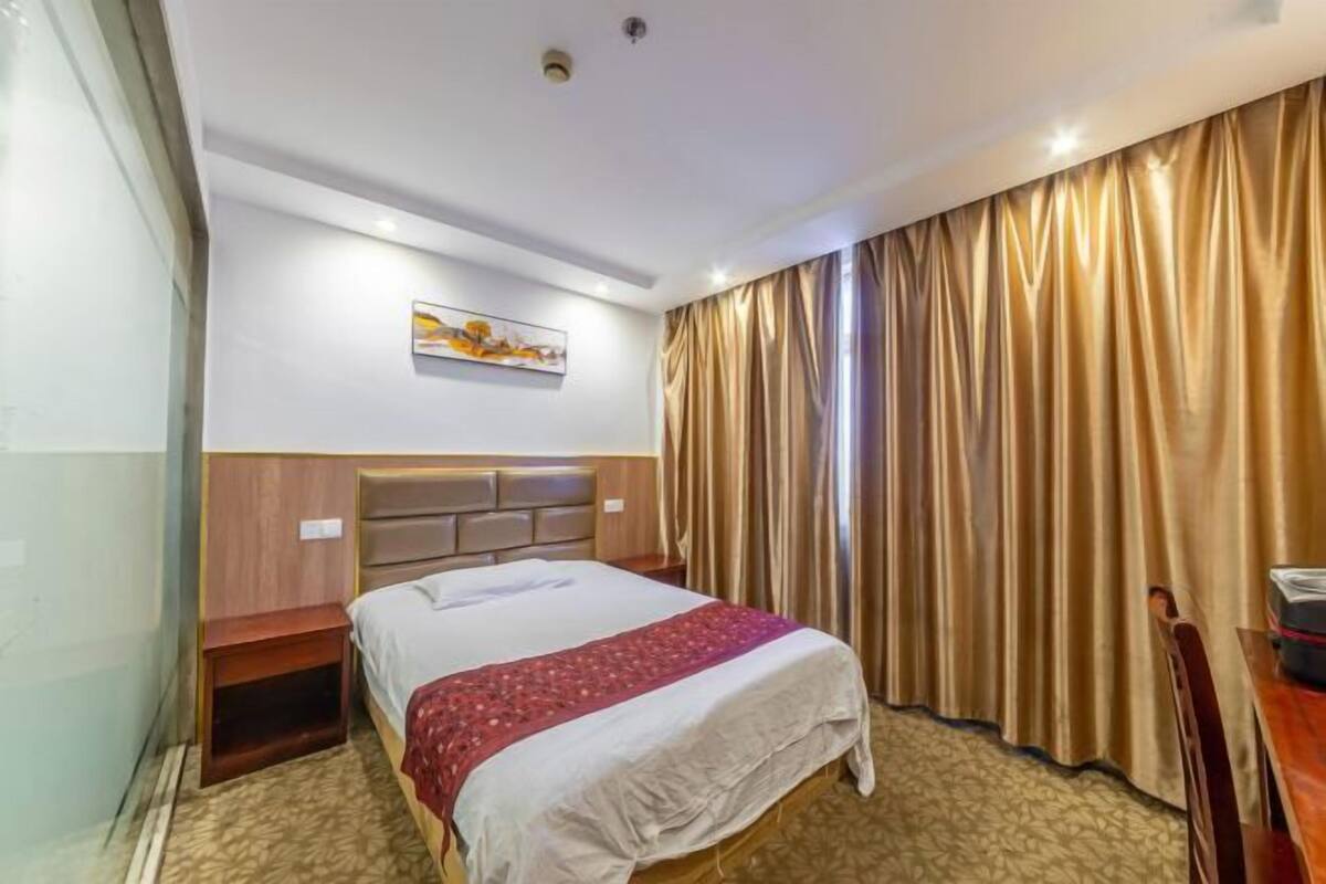 Una camera d'albergo con un letto, un comodino in legno, una testiera, un quadro appeso al muro e un'ampia finestra con tende.
