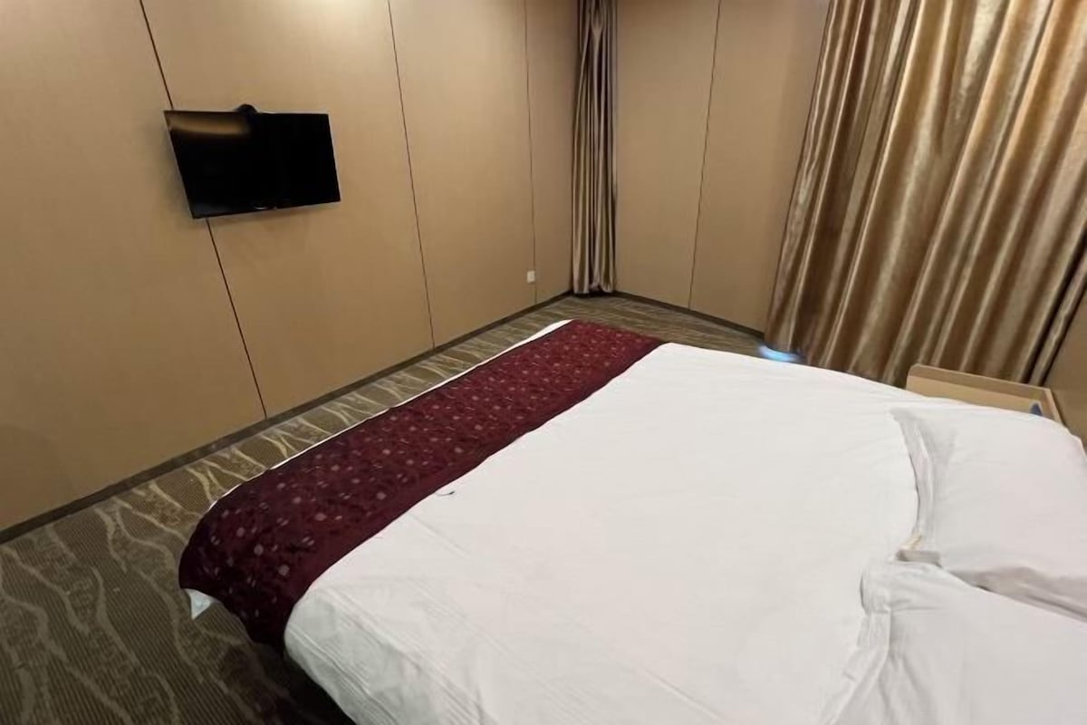 Ett hotellrum med en säng, en platt-TV som är monterad på väggen, ett mönstrat golv och ett fönster med gardiner.