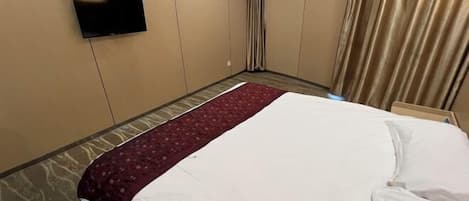 Una habitación de hotel con cama, televisor de pantalla plana montado en la pared, alfombra estampada y ventana con cortinas.