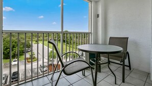 Un balcon avec une table ronde et deux chaises, une vue sur un parking et un ciel bleu dégagé.