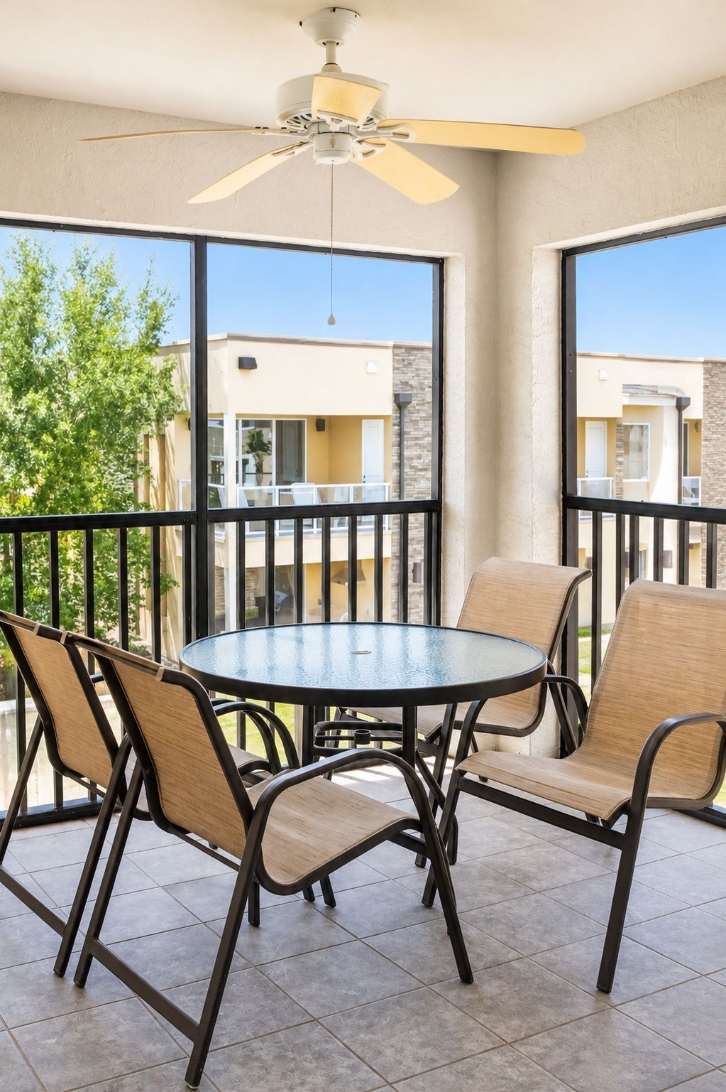 Een balkon met een ronde tafel en stoelen, een plafondventilator en uitzicht op omliggende gebouwen.