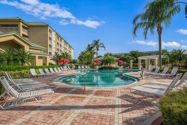 Un espace piscine doté de chaises longues et de parasols, entouré de palmiers et d’un bâtiment avec des balcons.