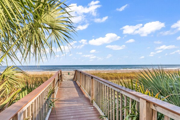 A wooden boardwalk leading to a beach with a clear sky and palm trees.