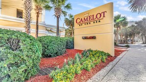 Un letrero de Camelot by the Sea con el aviso 'VACANCY' debajo, rodeado de jardines y palmeras.