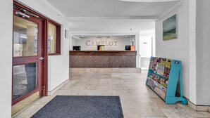 Zona de recepción con un cartel 'CAMELOT', un mostrador de recepción, un tablón de anuncios y una escalera que lleva a un nivel superior.