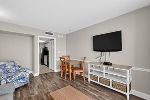 A compact living space with a sofa, a dining table with chairs, a TV mounted on the wall, and a kitchenette in the background.