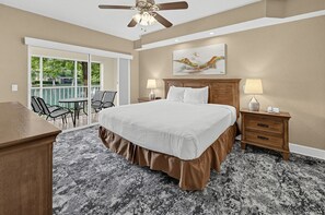 A hotel room with a large bed, a desk, a chair, a balcony with a view of trees, and a ceiling fan.