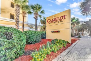 A sign for Camelot by the Sea with a 'VACANCY' sign below it, surrounded by landscaped gardens and palm trees.