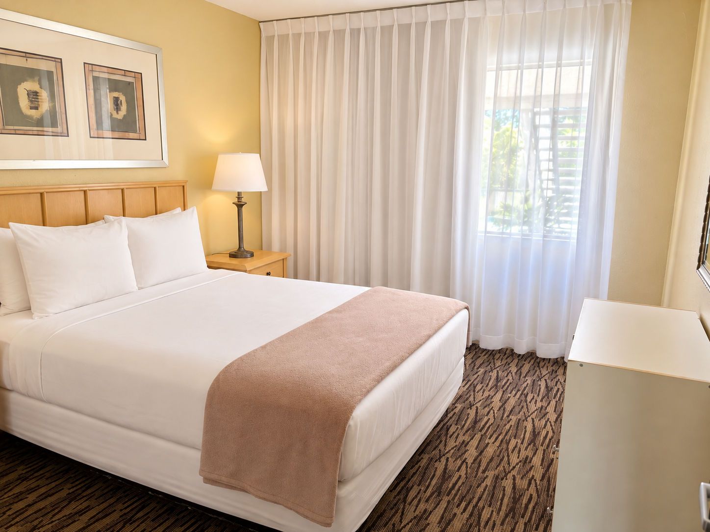 A hotel room with a bed, a nightstand, a lamp, a desk, and a window with curtains.