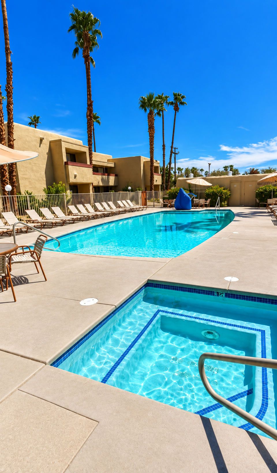 A swimming pool with lounge chairs and umbrellas, surrounded by palm trees and a building.