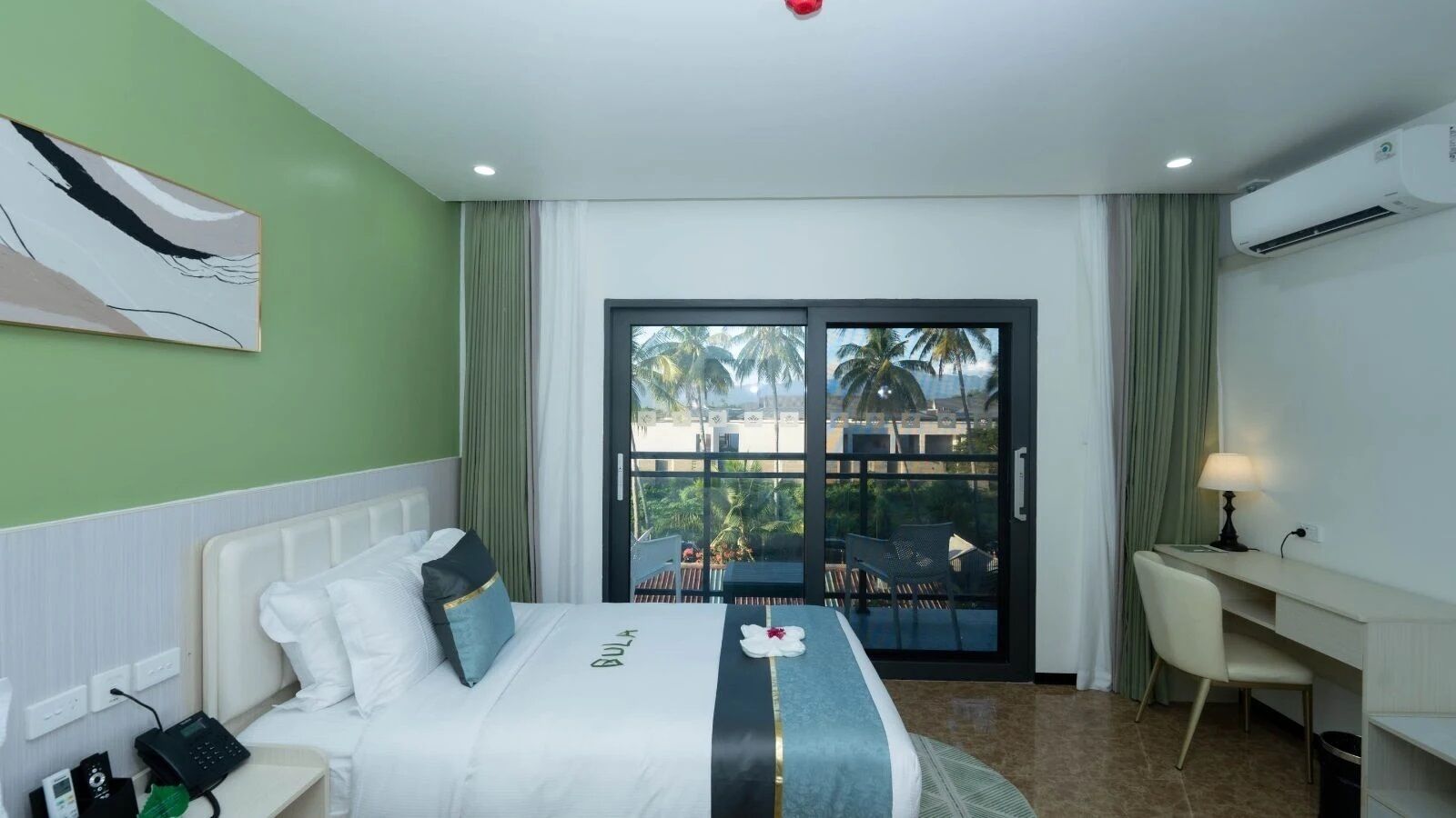 Ein Hotelzimmer mit Bett, Schreibtisch und Blick auf Palmen und einen Strand.