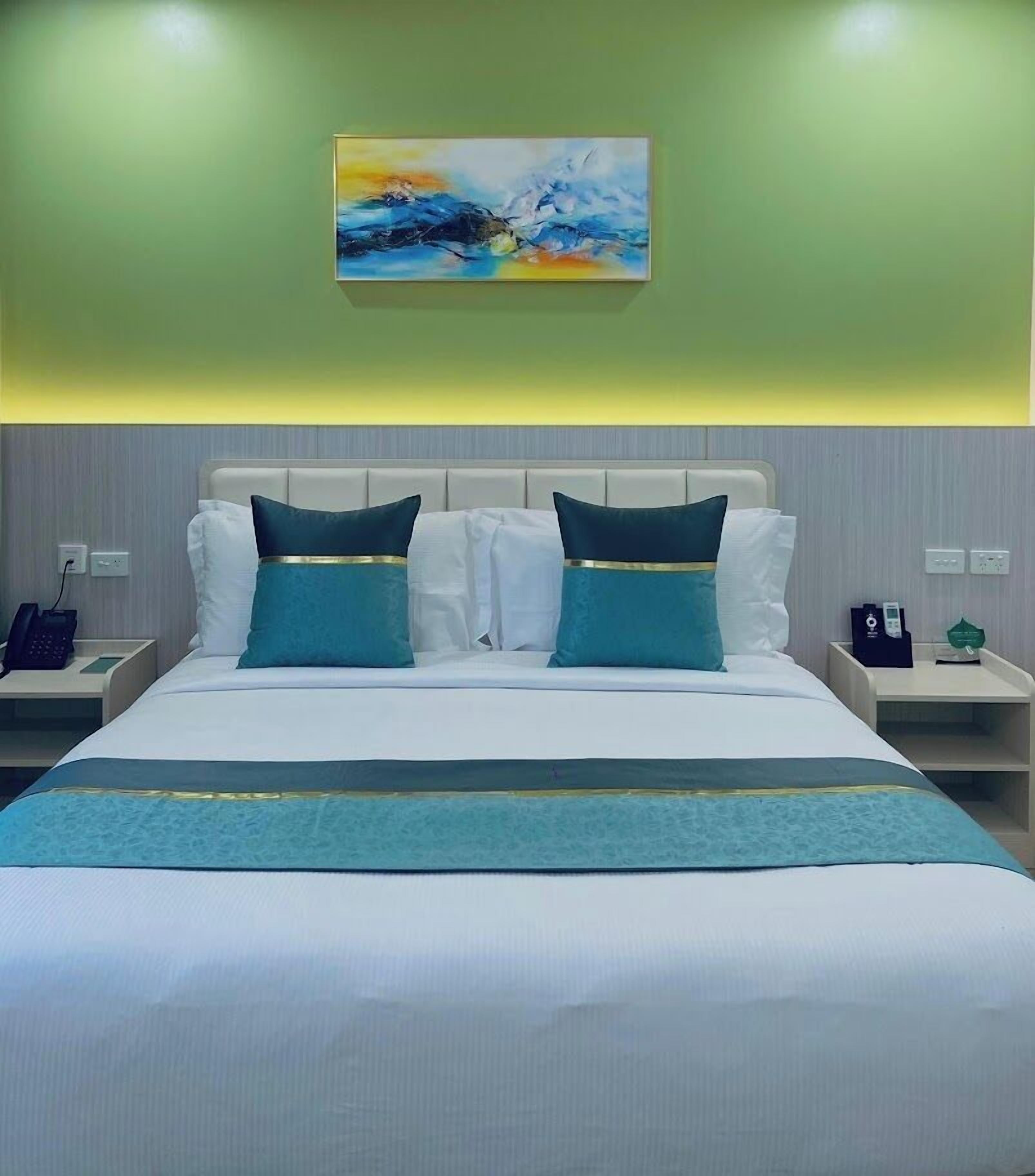 A hotel room with a large bed, two nightstands, a painting above the bed, and a green wall.