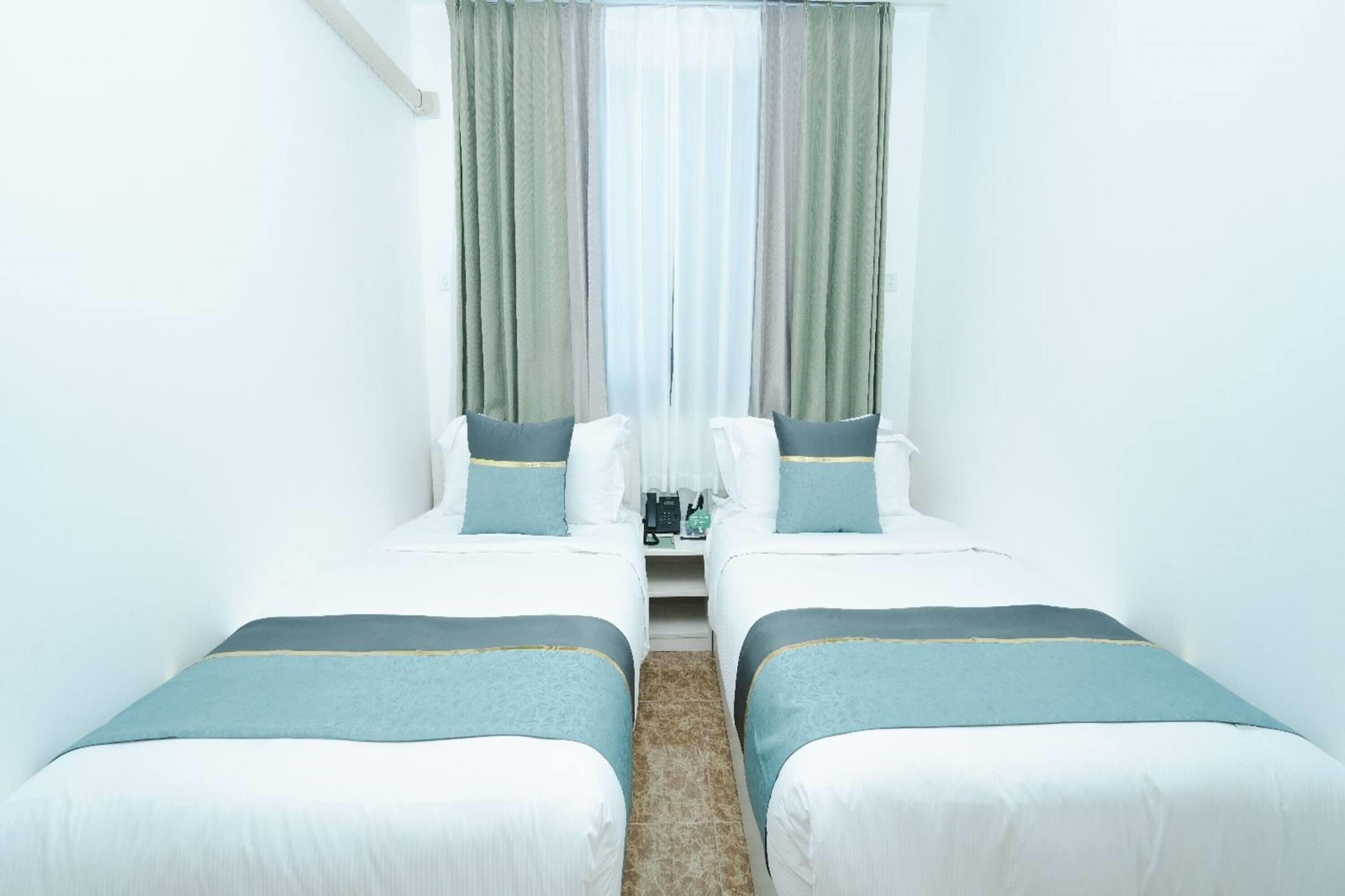 Une chambre d’hôtel avec deux lits, une fenêtre avec des rideaux légers et une petite table avec une télécommande.