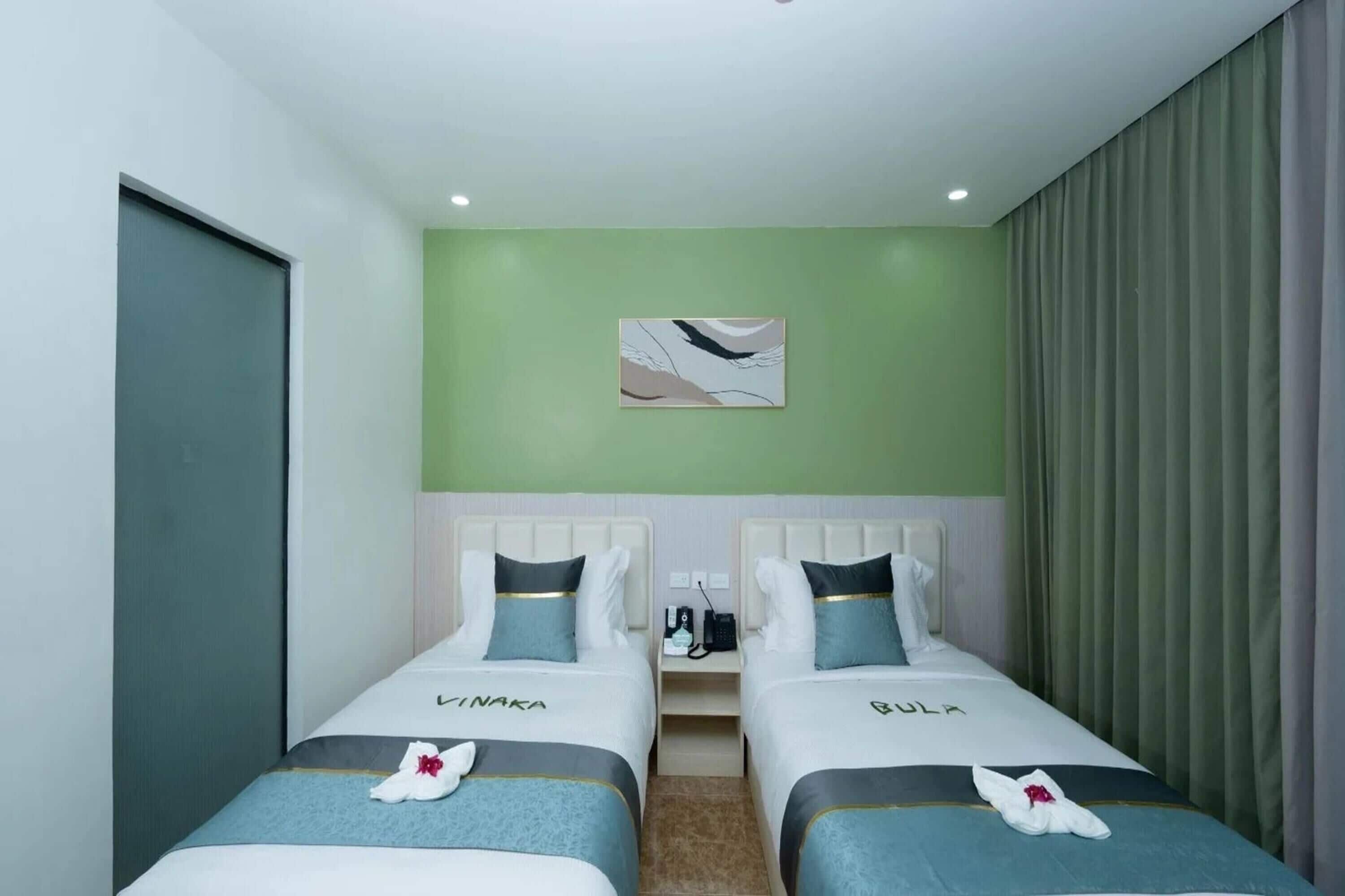 A hotel room with two beds, each with a name tag, a small table between them, and a painting on the wall.