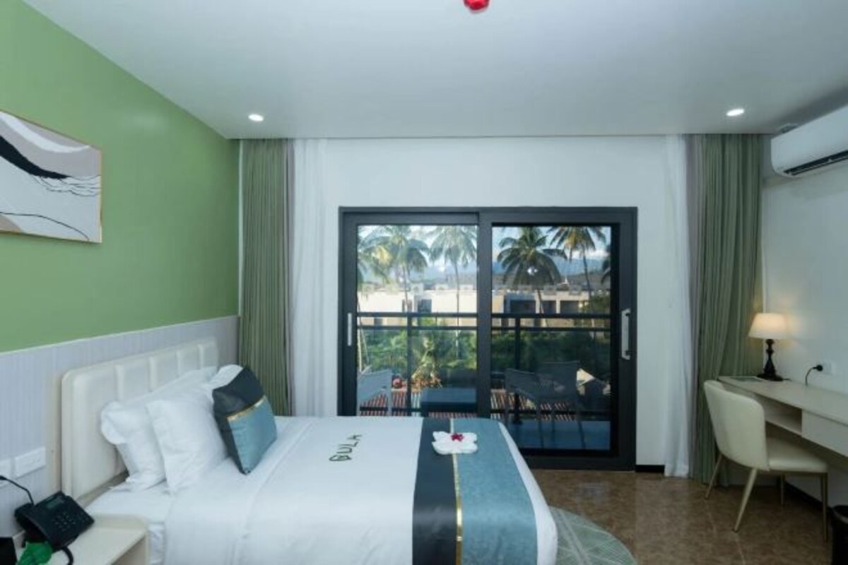A hotel room with a bed, a desk, and a view of palm trees through the glass door.