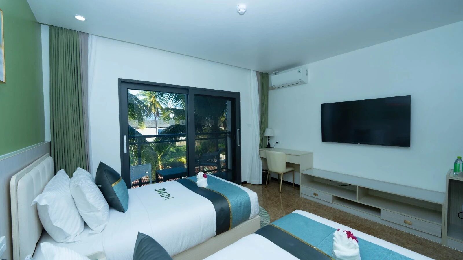 A hotel room with a bed, a desk, a TV, and a balcony with a view of palm trees.