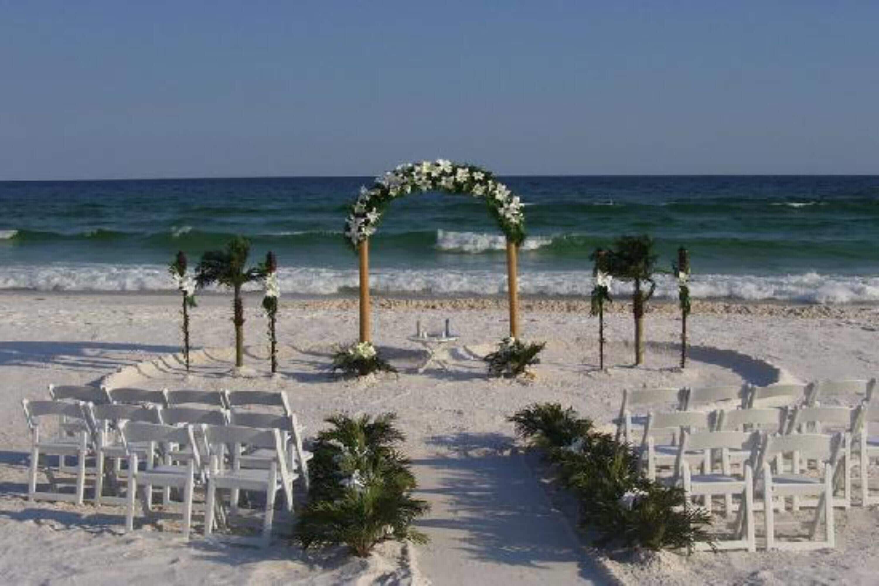 A beach wedding setup with white chairs and a floral arch.