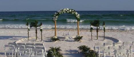 A beach wedding setup with white chairs and a floral arch.