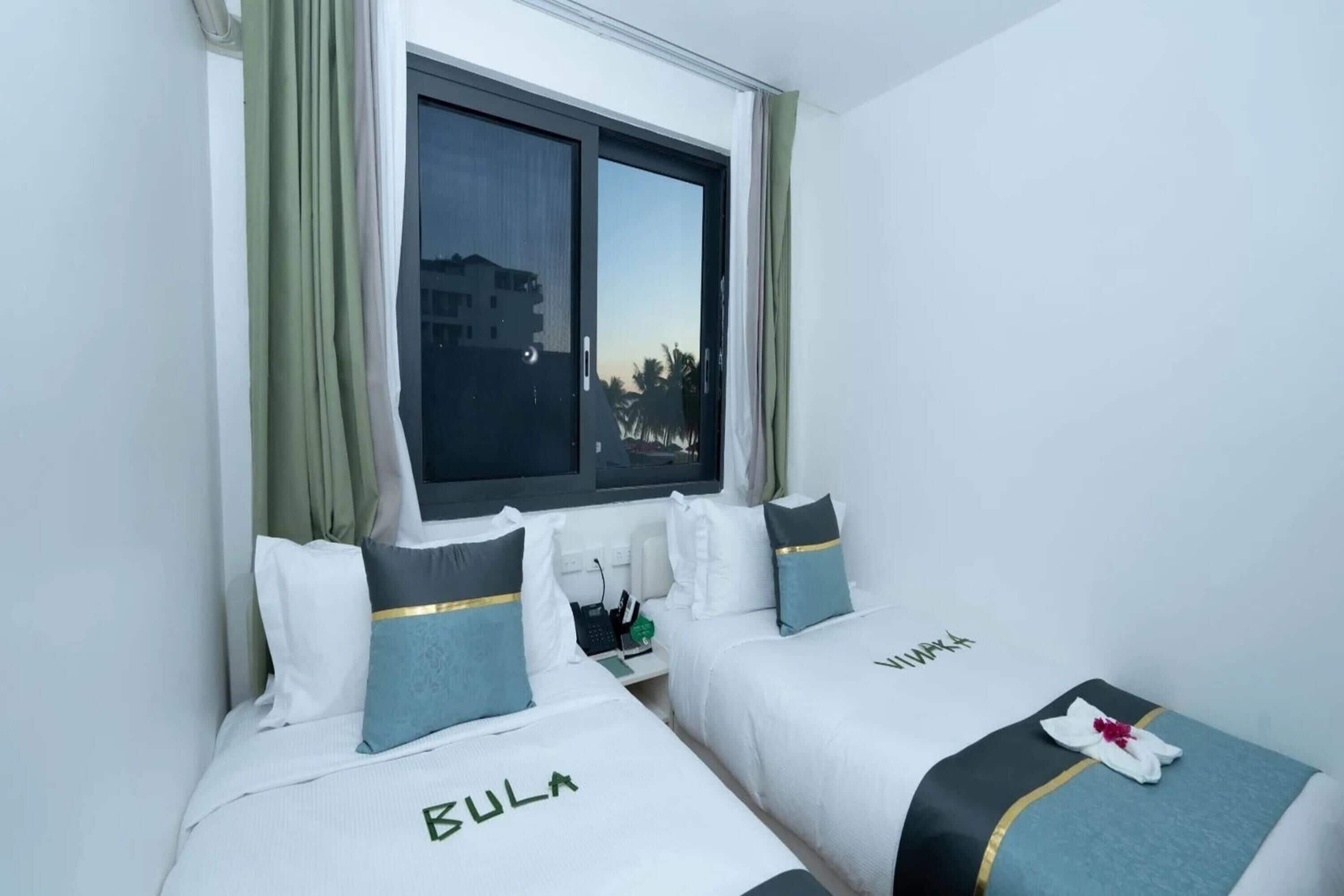 Habitación de hotel con dos camas, una ventana con cortinas y vistas a palmeras.