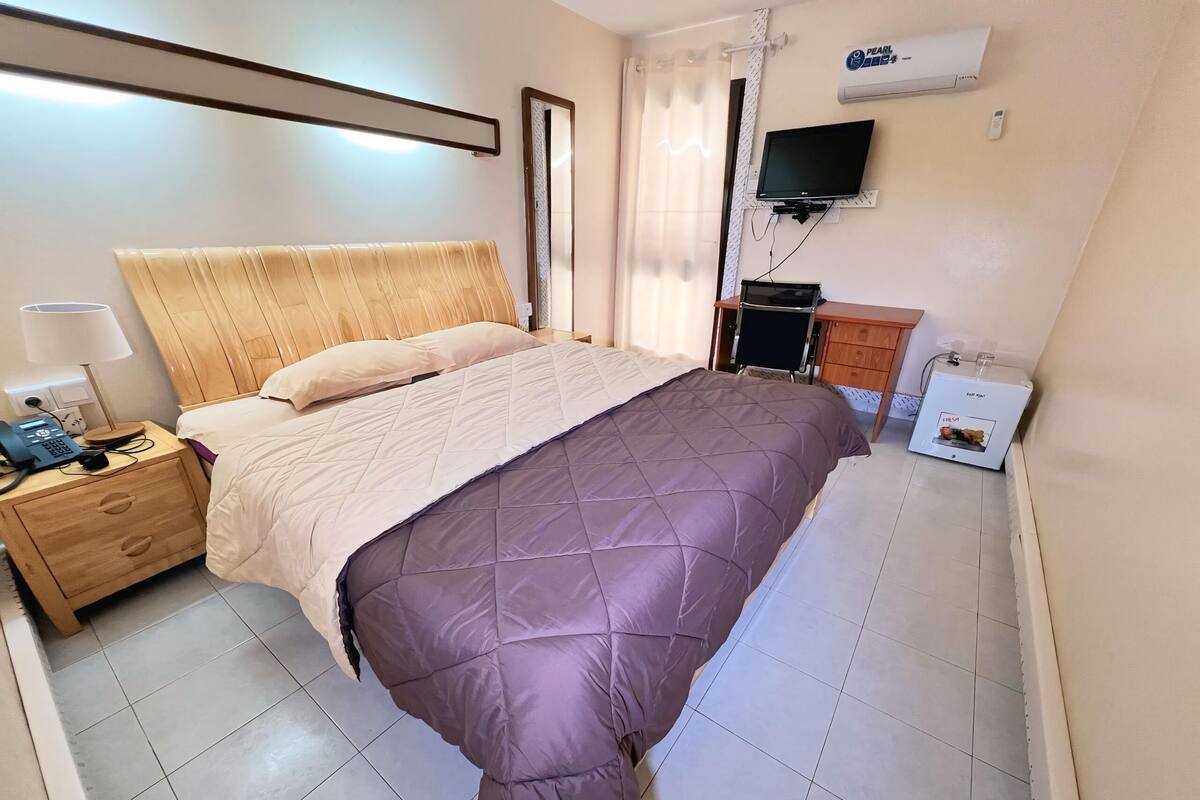 A bedroom with a wooden headboard, a bed with a purple quilt, a bedside table with a lamp, a TV mounted on the wall, and a small heater on the floor.