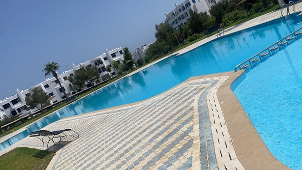 A swimming pool with a tiled deck, surrounded by white buildings and greenery.