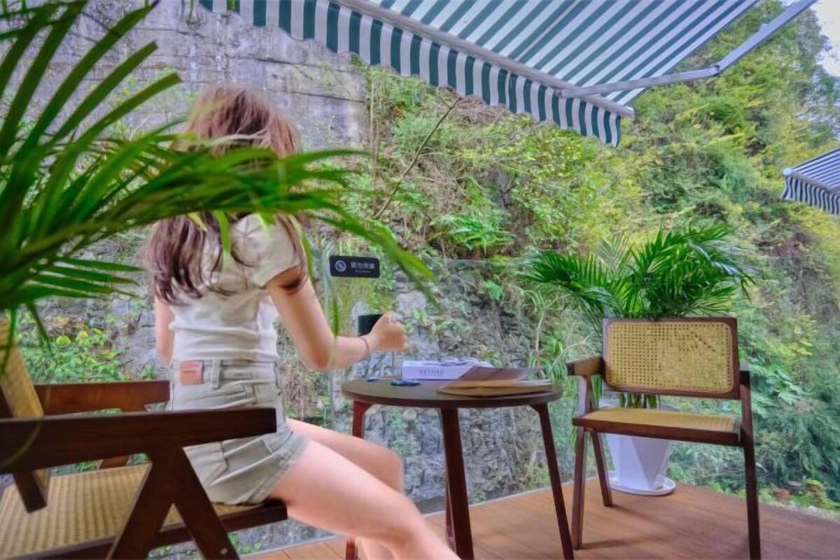 A person sitting at a table with a notebook and pen, surrounded by greenery and a stone wall.