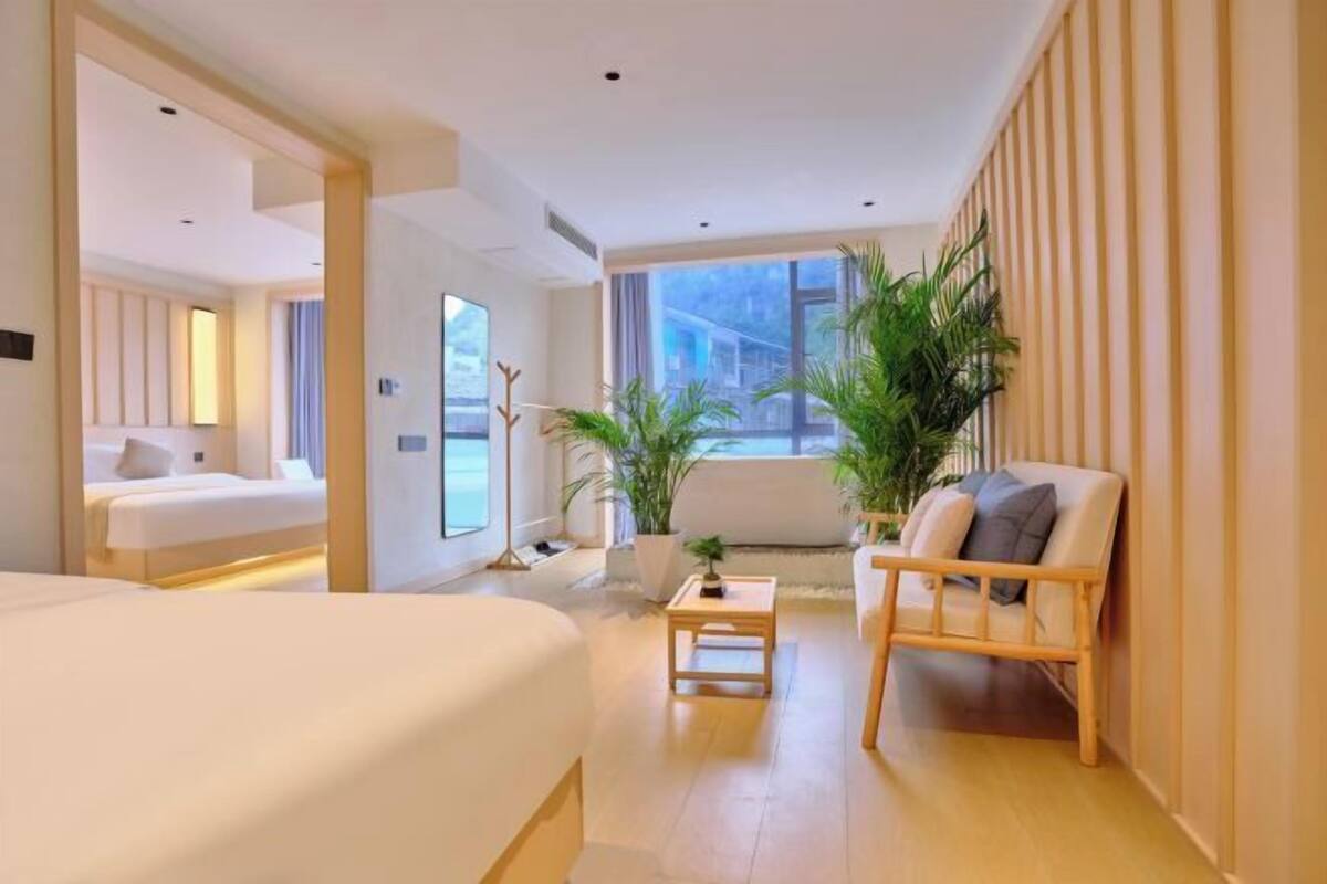 A modern hotel room with a large bed, a seating area with a chair and a small table, and large windows offering a view of the outside.