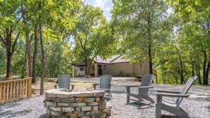Un área de estar al aire libre de estilo rústico, con sillas Adirondack y una chimenea de piedra, rodeada de árboles y una terraza de madera.