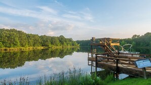 Un muelle de madera se adentra en un lago tranquilo con una orilla cubierta de bosques. Se observa una señalización en el muelle.