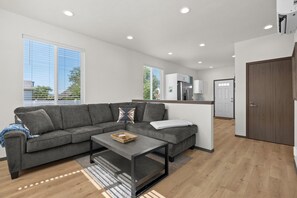 A modern living room with a grey sectional sofa, a coffee table, and a kitchen area with white cabinets and a refrigerator. - Clean 2 Bed Bungalow, Free Parking, Fast WiFi, Near GU (Spokane)