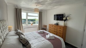 Una cama bien hecha con cabecera, un aparador de madera, un televisor en la pared y una ventana con cortinas.