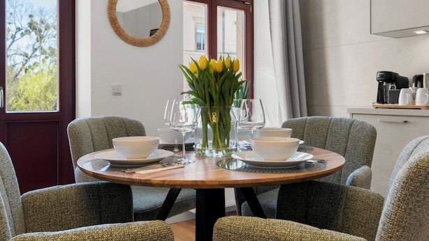 A dining area with a round table set for two, surrounded by upholstered chairs. A vase with yellow flowers is placed on the table. - ElbDreams Cozy Design Apartment (Dresden)