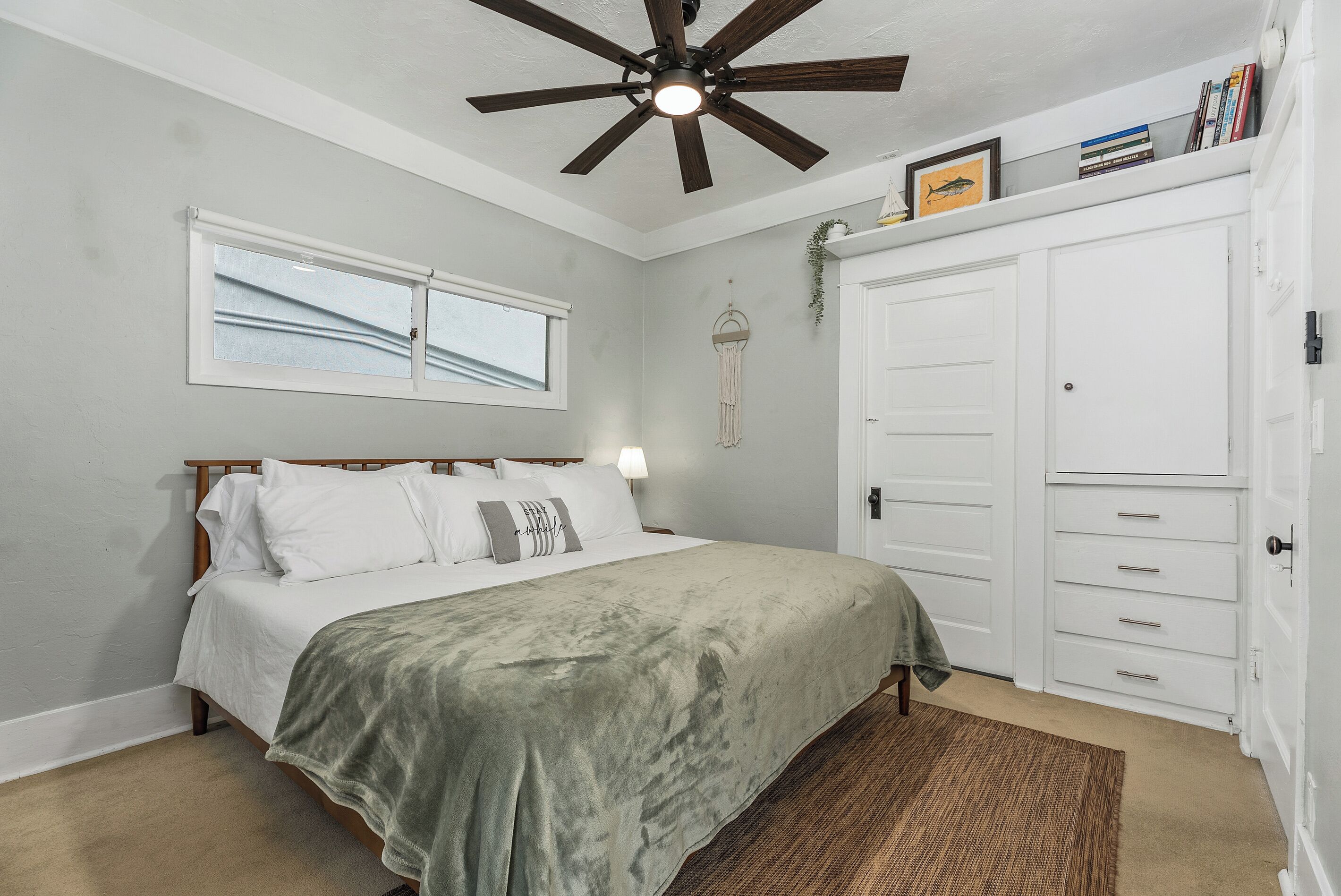 Un dormitorio con cama, ventilador de techo, ventana, armario y lámpara de noche.