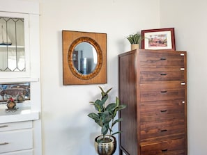 A room with a wooden cabinet, a mirror with a woven frame, a potted plant, and a framed picture on the wall.