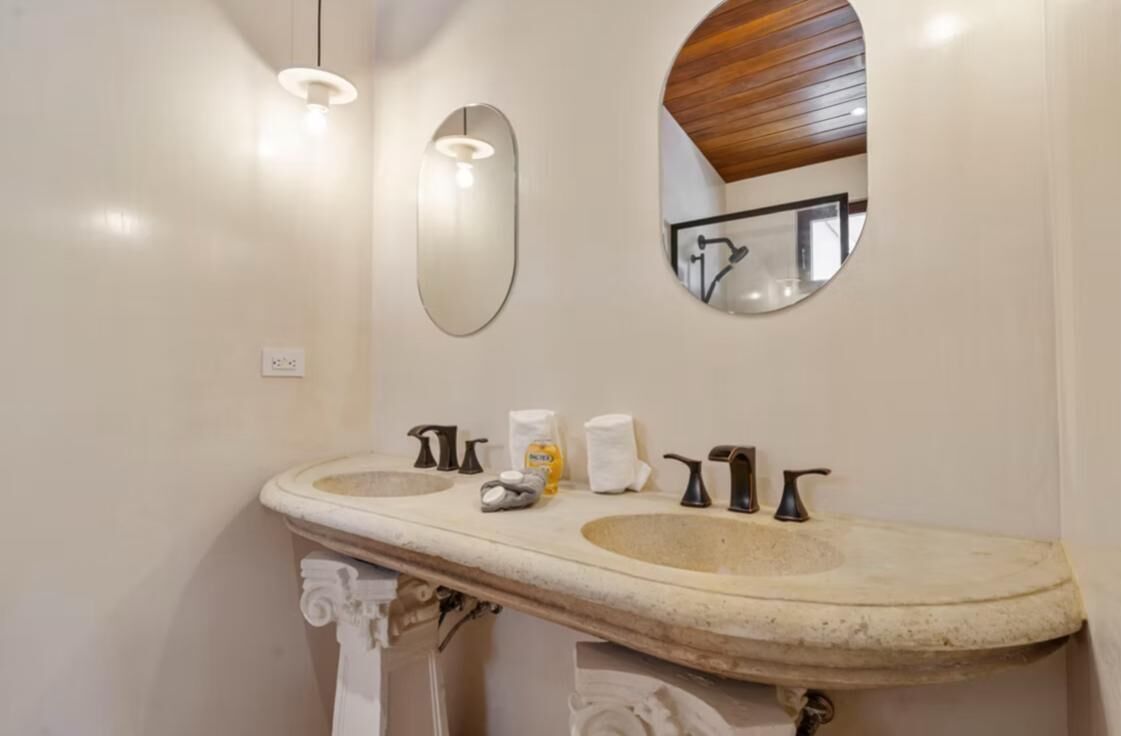 A bathroom with a double sink, two wall-mounted lights, a mirror, and a towel dispenser.