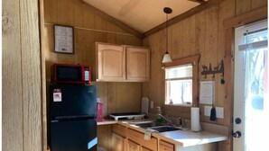 A compact kitchen with wooden cabinets, a black refrigerator, a microwave, a sink, and a window with blinds. - Pop's Place on the Rapidan River. (Rochelle)