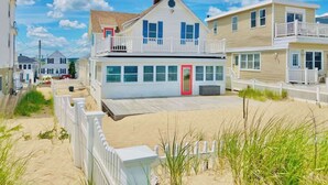 A beachfront house with a red door, white railings, and a sandy walkway.
